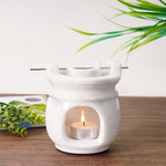 White ceramic oil burner with a lit tealight candle on a wooden surface with green plants in the background.