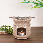 Ceramic oil burner with a lit candle on a wooden surface with greenery in the background.
