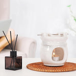White ceramic oil burner with a lit candle on a woven mat, next to a black diffuser bottle.