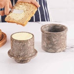 A natural marble butter crock and lid on a white surface, with a person spreading butter on a slice of bread using a knife.