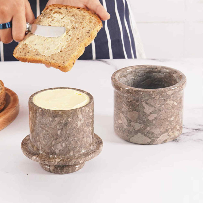 A natural marble butter crock and lid on a white surface, with a person spreading butter on a slice of bread using a knife.