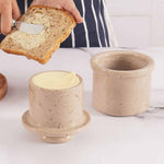 A natural marble butter crock and lid on a white surface, with a person spreading butter on a slice of bread using a knife.