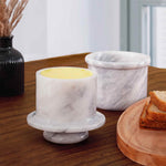 A natural marble butter crock and lid on a white surface, with a person spreading butter on a slice of bread using a knife.